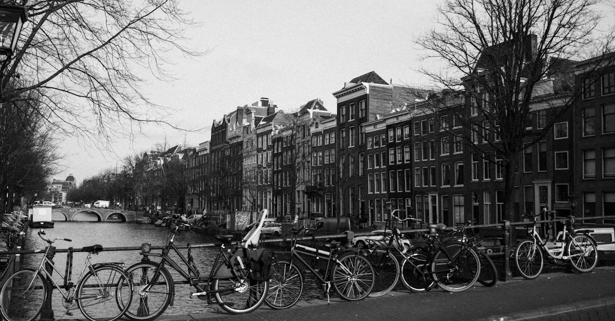 black-and-white-of-various-bicycles-parked-on-paved-bridge-over-rippling-canal-near-aged-typical-res
