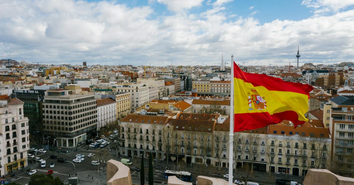 drone-view-of-spanish-city-with-aged-buildings-and-national-flag-under-cloudy-blue-sky