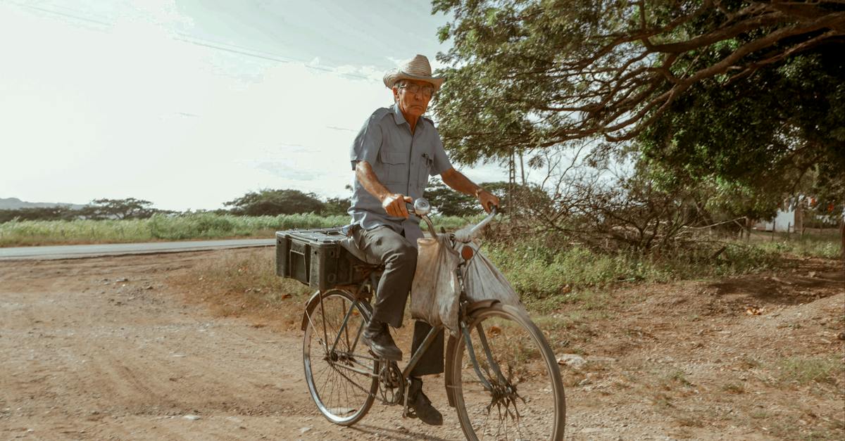 elderly-male-in-hat-riding-bike-on-dry-roadway-and-looking-at-camera-under-cloudy-sky