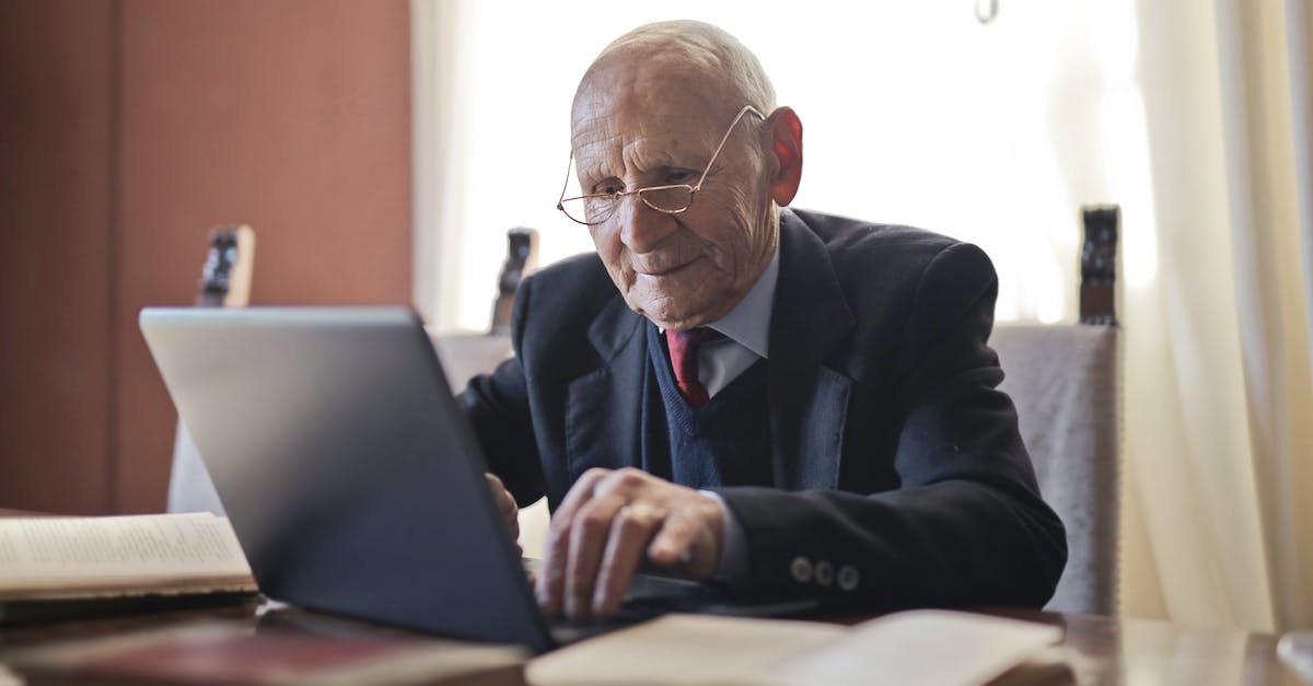 focused-elderly-man-in-formal-black-suit-and-eyeglasses-using-laptop-while-sitting-at-wooden-table-w-1