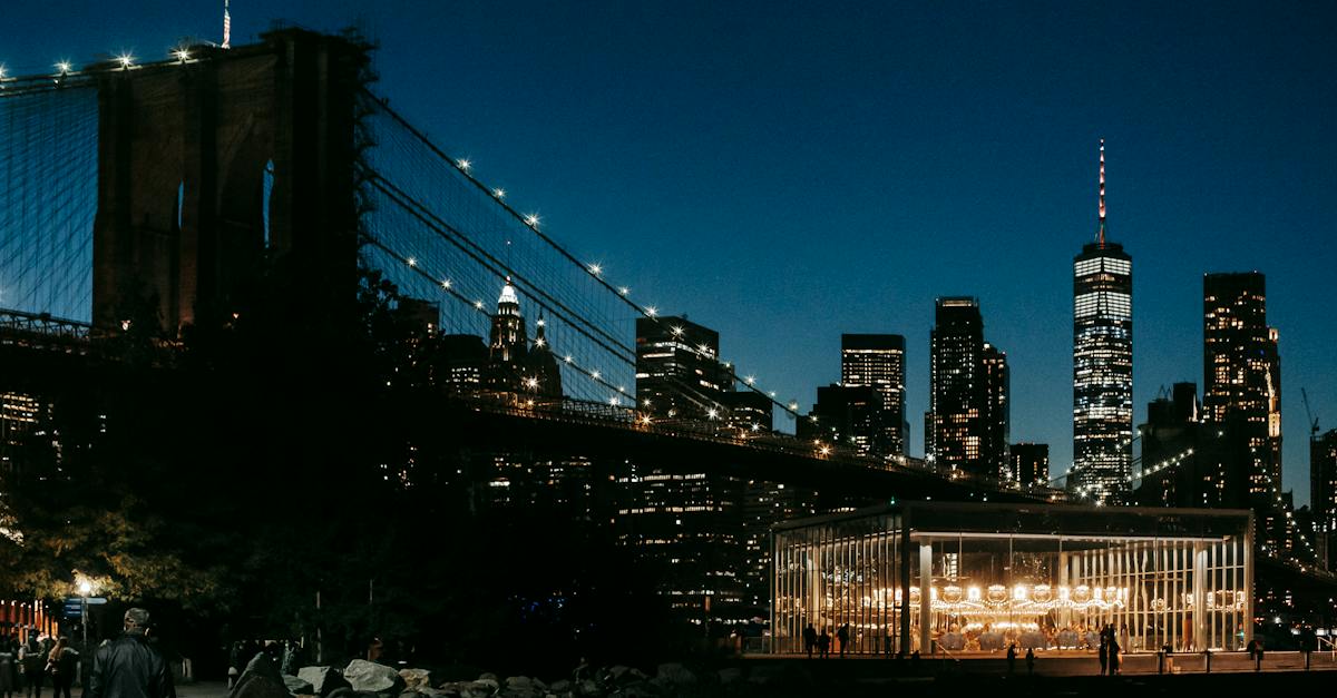 glowing-city-lights-on-high-rise-buildings-and-suspension-bridge-over-river-with-sidewalk-on-embankm