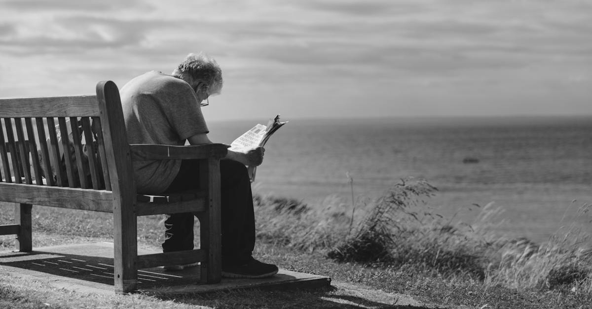 grayscale-photo-of-man-sitting-on-brown-wooden-bench-reading-news-paper-during-day-time