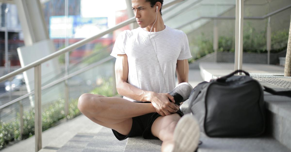 young-ethnic-athlete-in-earbuds-stretching-body-while-sitting-on-stairs-at-entrance-of-contemporary