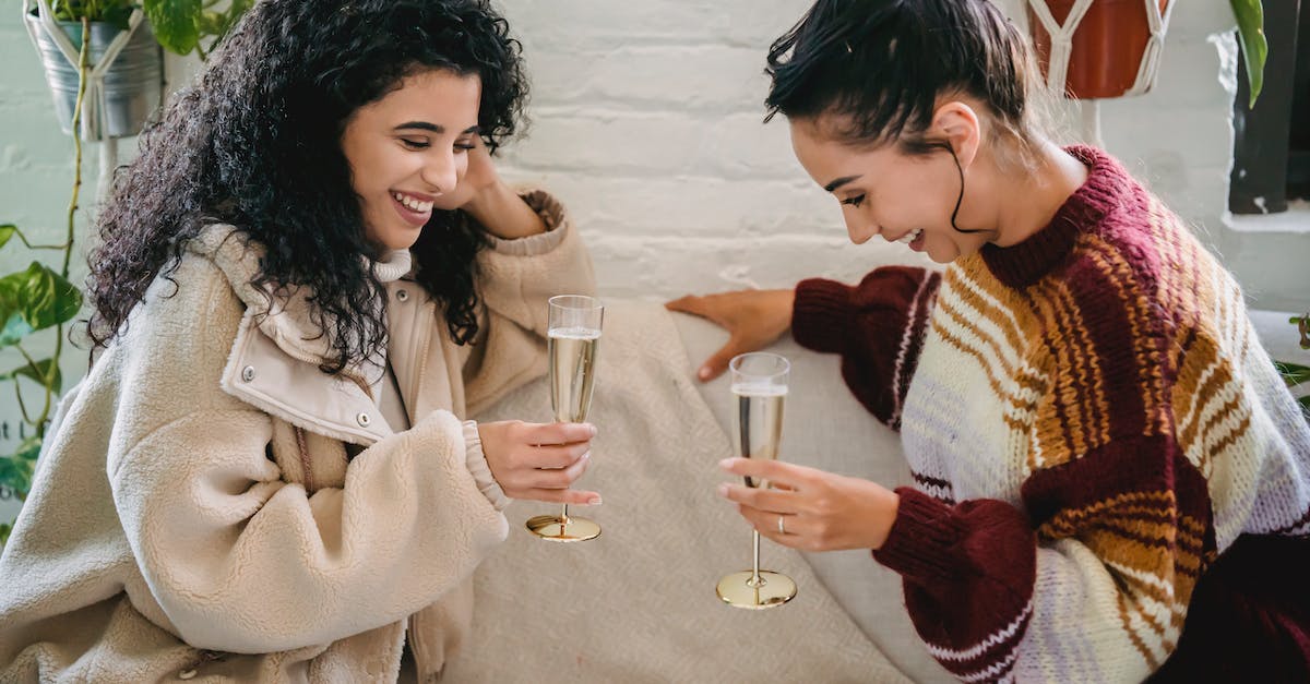 young-happy-hispanic-female-friends-with-glasses-of-champagne-having-fun-sitting-on-sofa-and-celebra