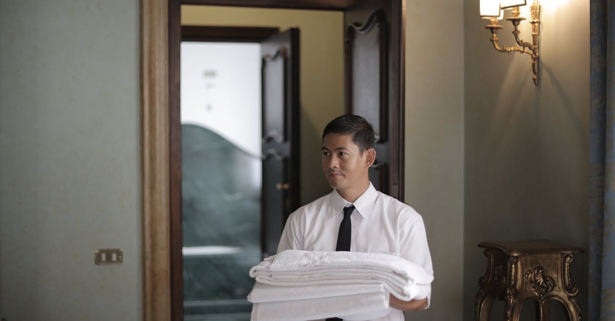 young-male-housekeeper-carrying-stack-of-white-bed-sheets-while-entering-bedroom-in-hotel-1